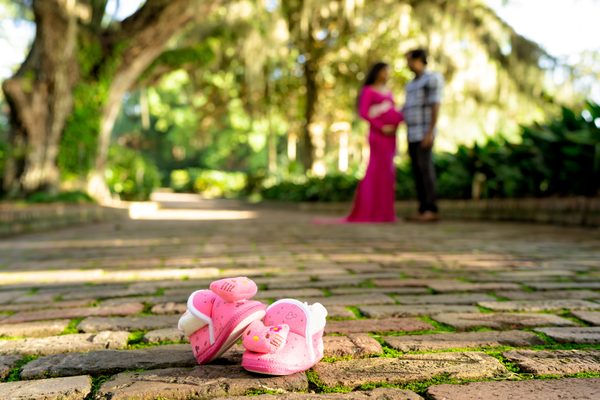 Expectant couple on a mossy brick terrace under live oaks, with pink baby shoes in the foreground, at Maclay Gardens