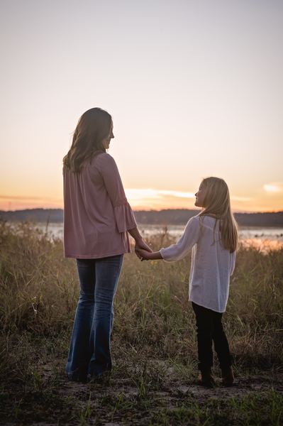 Mother and daughter seen from behind, holding hands in tall grass and facing Lake Jackson at sunset at Millers Landing